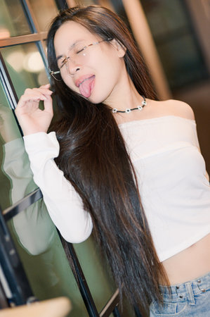 A joyful young woman in a casual outfit playfully poses while sticking out her tongue. Her long hair cascades down, and she wears stylish glasses, exuding a carefree and trendy vibe.の写真素材