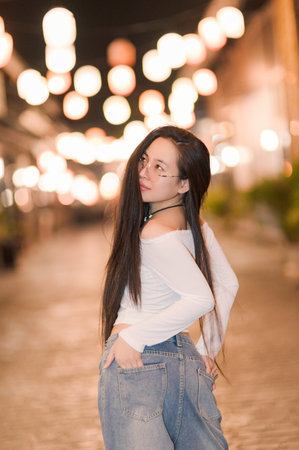 A young woman with long hair poses confidently against a softly lit backdrop. The warm street lights create a serene atmosphere, showcasing her stylish outfit.の写真素材