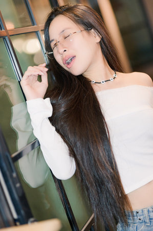 A young woman with long, flowing hair exudes elegance and joy in a modern indoor setting. Her confident pose and stylish glasses create a captivating portrait.の写真素材