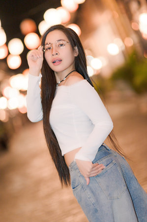 A stylish young woman poses confidently outdoors at night, surrounded by softly glowing lights. Her long hair and casual fashion complement the serene atmosphere.の写真素材