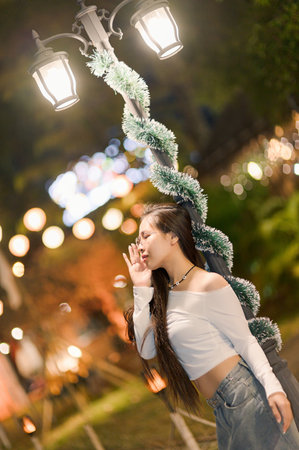A young woman poses playfully near a beautifully decorated street lamp at night. The festive atmosphere is enhanced by enchanting lights and greenery.の写真素材