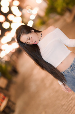 A young woman with long hair poses casually in an urban night scene. The background features bright lights, creating a soft bokeh effect, highlighting her relaxed enjoyment.の写真素材