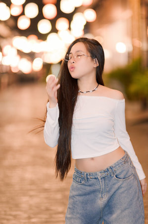 A young woman showcases her playful spirit as she poses confidently in a vibrant street setting illuminated by soft lights. Her casual outfit adds a trendy touch to the joyful atmosphere.の写真素材