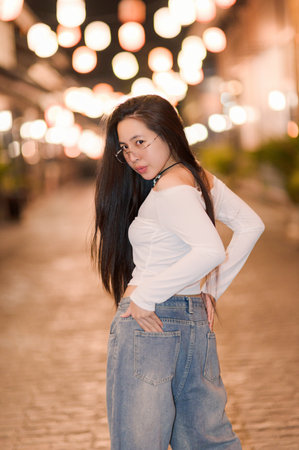 A confident young woman poses in a trendy outfit on a charming urban street illuminated by lanterns, creating a magical night atmosphere.の写真素材