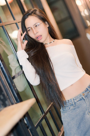 A stylish young woman poses confidently against a glass wall, showcasing her long hair and trendy outfit. The image captures modern fashion and intimate beauty.の写真素材