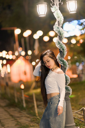A young woman poses gracefully under a beautifully lit street lamp during a lively nighttime festival. The scene captures her relaxed expression and stylish outfit amidst a backdrop of twinkling lights and festive decorations.の写真素材