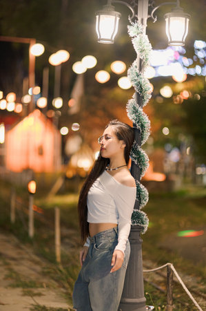 A young woman poses gracefully against a street lamp adorned with festive decorations. The scene captures a serene and joyful moment in an illuminated outdoor environment.の写真素材