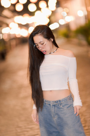A young woman with long hair stands in a softly lit street at night, exuding confidence and style. The glowing lights and casual attire create a charming atmosphere.の写真素材