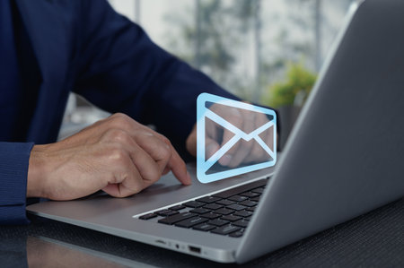 A person typing on a laptop in a modern workspace with a translucent email icon above the keyboard, symbolizing digital communication and productivity.の写真素材