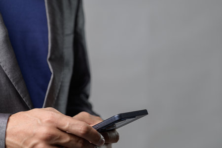 A man in a suit is holding a smartphone with a gray background. The image captures modern technology in a professional yet casual setting, perfect for business-related themes.の写真素材
