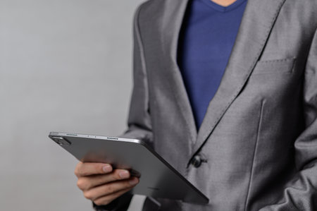 A business professional holds a modern tablet in one hand, dressed in a stylish gray suit. This close-up captures confidence and innovation in a corporate setting.の写真素材