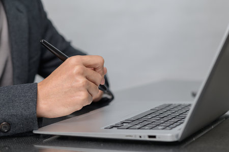 A close-up image of a hand holding a pen, writing on a laptop in a modern workspace. This scene captures the essence of productivity and professionalism in a corporate environment.の写真素材