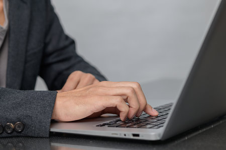 A close-up image of a person's hands typing on a laptop keyboard, set against a soft gray background. The individual is dressed in professional attire, highlighting a modern workspace.の写真素材