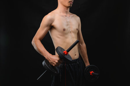 A focused young man stands with dumbbells, showcasing determination to improve strength and health. The dark background emphasizes his physique and commitment to fitness.の写真素材