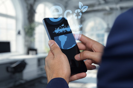 A person uses a smartphone to engage with eco friendly technology. Digital graphics highlight a commitment to sustainability in a modern workspace setting.の写真素材