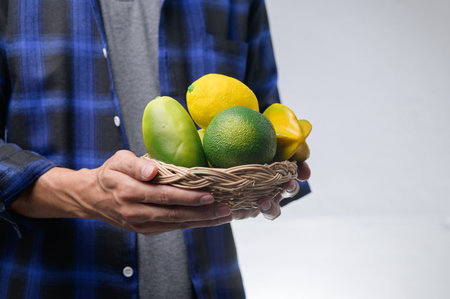 A person in a plaid shirt holds a basket filled with bright and fresh organic fruits. The arrangement includes mango, lemon, and lime, showcasing a vibrant and healthy selection.の写真素材