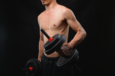 A young man showcases physical strength by lifting dumbbells against a black background, emphasizing fitness and health. This image captures the essence of determination in an active lifestyle.の写真素材