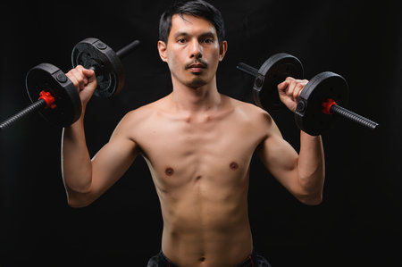 A determined man lifts dumbbells in a studio setting, showcasing strength and dedication to fitness. The dark background emphasizes his physique and focus on health.の写真素材