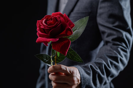 A sophisticated man holds a vibrant red rose in a dark setting, symbolizing love and romance. The elegant gesture evokes emotions of passion and beauty.の写真素材