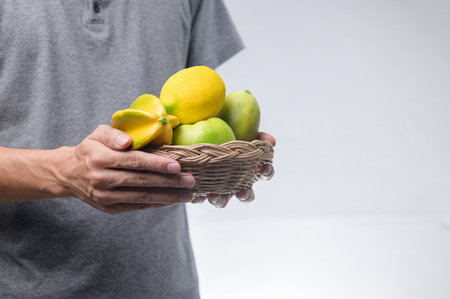 A woven basket filled with vibrant fresh fruits including lemons, limes, and mangoes, held by a person's hand against a simple backdrop, showcasing healthy living.の写真素材