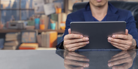 A businessman attentively uses a tablet in a stylish office setting. The image captures the reflection on the desk surface, highlighting modern technology and a productive environment.の写真素材