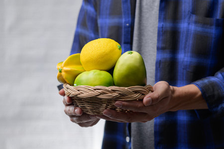 A person holds a woven basket filled with fresh fruits, including lemon and green apples. The blue plaid shirt adds a rustic touch to this colorful harvest scene.の写真素材