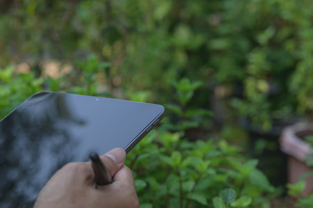 A close-up view of a hand holding a sleek tablet in a vibrant green garden. The lush foliage provides a serene backdrop, perfect for relaxation and creativity.の写真素材