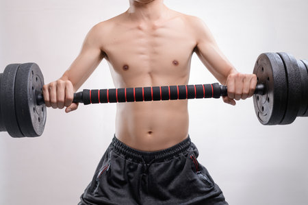 A young man demonstrates strength training by lifting a barbell in a fitness-focused environment. The image captures his toned physique and dedication to health.の写真素材