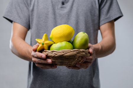 A person holds a woven basket filled with vibrant tropical fruits, showcasing a lemon, lime, mango, and starfruit. Perfect for healthy lifestyle imagery.の写真素材
