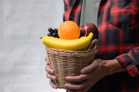 A close-up view of a person holding a wicker basket filled with fresh, colorful fruits including an orange, banana, apple, and grapes, symbolizing healthy living.の写真素材