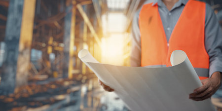 A construction worker in an orange safety vest holds a blueprint in an industrial setting. The warm sunlight creates a dynamic atmosphere ideal for project planning.の写真素材