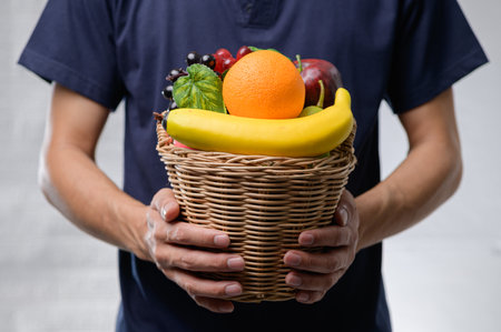 A person holds a wicker basket filled with assorted fresh fruits, including bananas, oranges, and grapes, showcasing healthy lifestyle choices against a neutral background.の写真素材