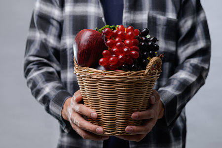 A close-up of a person holding a woven basket filled with fresh fruits, including grapes and an apple, dressed in a plaid shirt against a neutral background.の写真素材