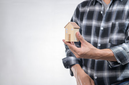 A man in a plaid shirt holds a small house model, symbolizing real estate investment and property ownership. This image captures the essence of housing market dynamics.の写真素材