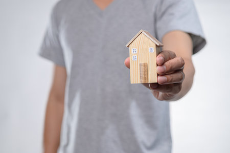 A hand presents a small wooden model house, symbolizing the essence of real estate investment and home ownership in a modern, minimalistic setting.の写真素材