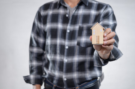 A person wearing a classic plaid flannel shirt holds a small wooden model of a house, symbolizing concepts of home, design, and investment in real estate.の写真素材