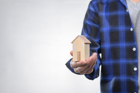 A person holds a miniature wooden house in one hand while wearing a casual blue checkered shirt. This image symbolizes home, security, and investment.の写真素材