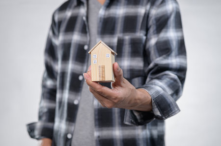 A person in a stylish flannel shirt holds a small wooden house model, symbolizing homeownership and property investment. The image captures the essence of creativity and dreams.の写真素材