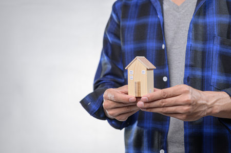A person holds a small wooden house model, symbolizing home ownership and potential investment, dressed in a casual blue flannel shirt, against a soft background.の写真素材
