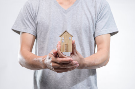 Individual in gray shirt gently holds a small wooden house model, symbolizing home ownership and real estate investment, against a clean white background.の写真素材