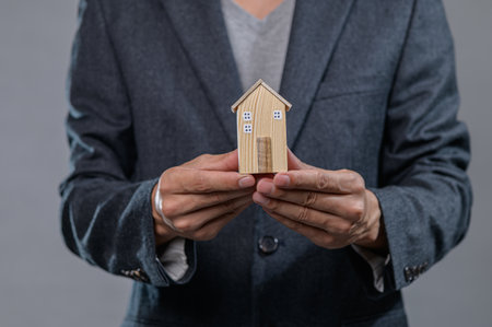 A businessman displays a small wooden model house, symbolizing real estate investment and property ownership. The elegant setting highlights professionalism and business acumen.の写真素材