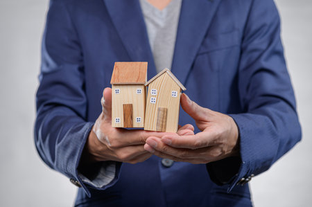 A close-up of hands holding two miniature wooden houses, representing concepts in real estate, investment, and home ownership. Ideal for business and finance themes.の写真素材