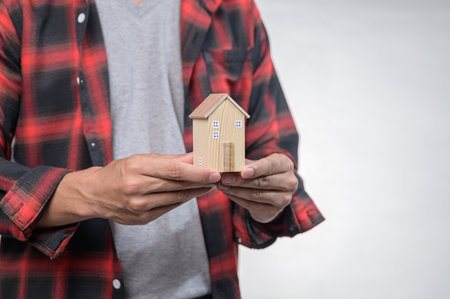 A person in a plaid shirt holds a small wooden house model, symbolizing homeownership and dreams. This image represents design, investment, and lifestyle choices.の写真素材