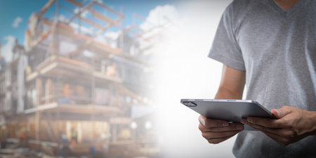 A construction worker uses a tablet to manage tasks on a busy site. This image represents the integration of technology in modern construction business practices, enhancing efficiency and collaboration.の写真素材