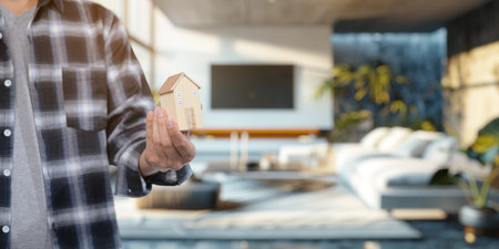 Explore the concept of buying and selling residential properties in a modern living room setting, featuring a person holding a model house, signifying investment and transaction.の写真素材