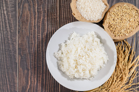 This image showcases a plate of fresh cooked white rice set on a rustic wooden table, accompanied by raw ingredients including rice grains and paddy. Ideal for culinary themes.の写真素材
