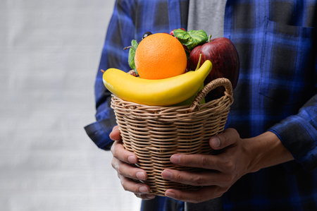 A person holds a woven basket filled with vibrant fresh fruits including an orange, banana, and apple. Perfect for promoting healthy eating and lifestyle.の写真素材