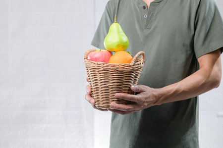 A person holds a basket filled with fresh fruits, including an apple, orange, and a pear, symbolizing health and nutrition in an indoor setting.の写真素材