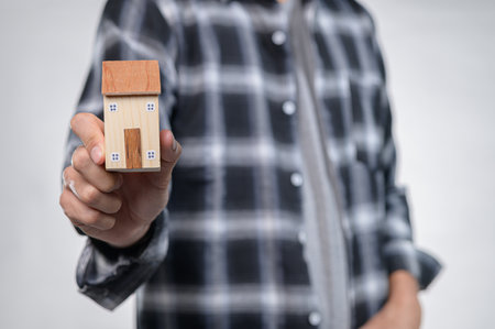 A person presents a small wooden house model, symbolizing home and comfort. The casual attire pairs well with the decorative element, representing lifestyle choices.の写真素材