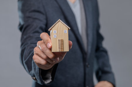 A businessman proudly presents a small wooden house model, symbolizing his commitment to the real estate market and investment opportunities.の写真素材